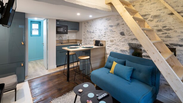 A modern living space with a blue sofa, wooden ladder, kitchen area, and a staircase.