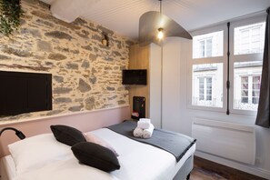 A bedroom with a stone wall, a bed with pillows, a TV, a radiator, and a window with a view of buildings. - Le Pink - Studio tout confort (Rennes)