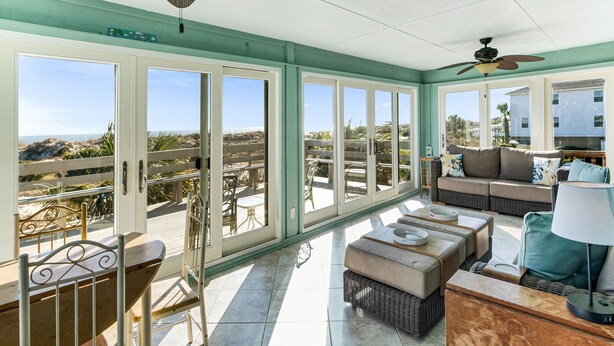 A spacious sunroom with large sliding glass doors, a comfortable seating area with a sofa and ottomans, and a dining table. - Oceanview Retreat | Just Steps from the Beach (Tybee Island)