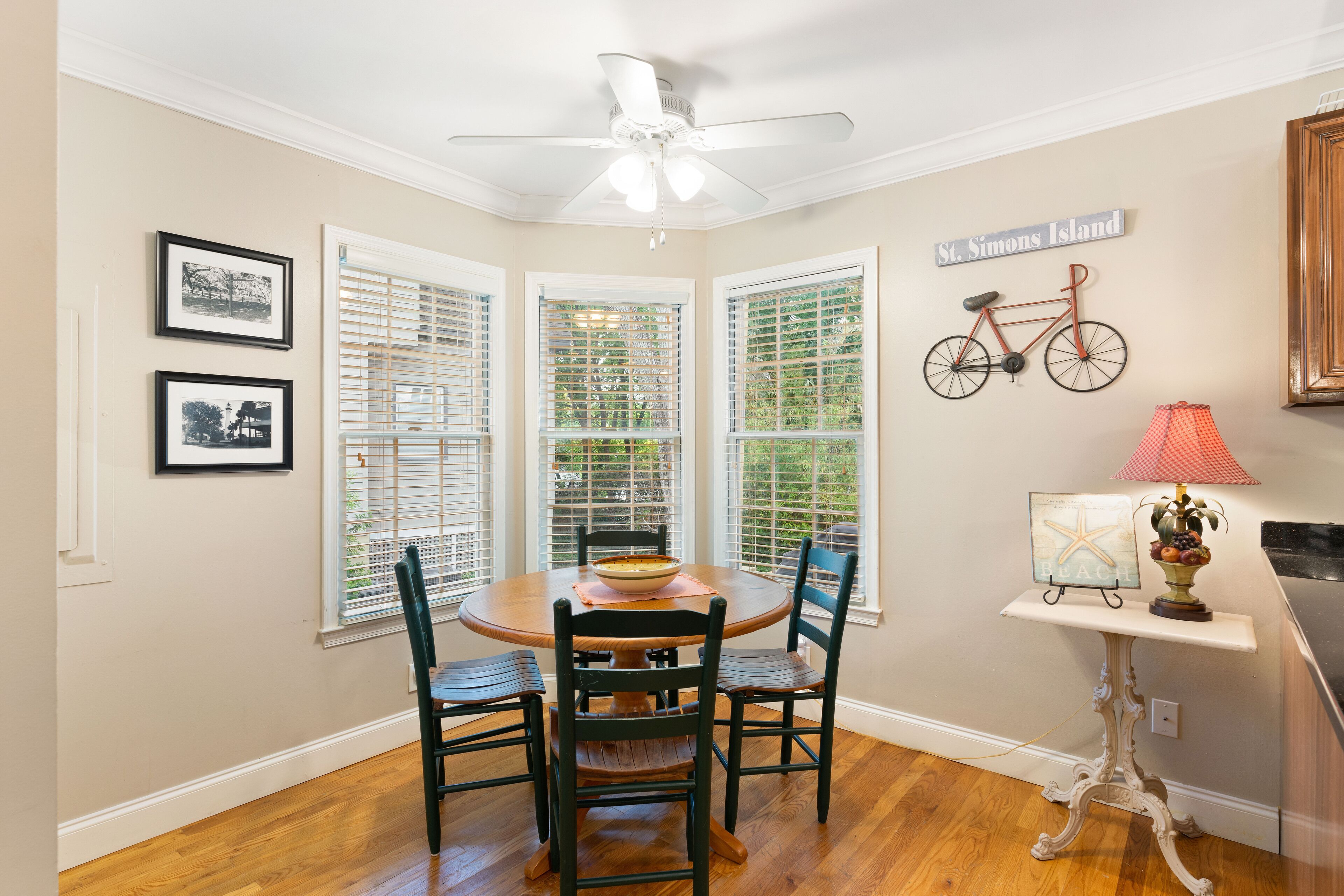 Uma sala de jantar com uma mesa redonda, cadeiras e um ventilador de teto. Há quadros nas paredes e uma placa com a inscrição 'St. Simons Island'.