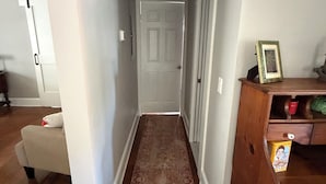 En smal hall med en mönstrad matta, en vit dörr längst ner och en trähylla med en bilderam och en låda på höger sida.