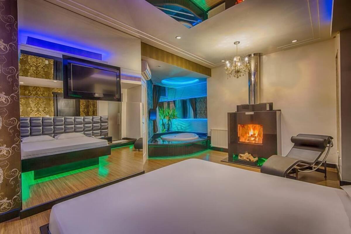 A modern hotel room with a bed, a TV, a fireplace, and a jacuzzi.