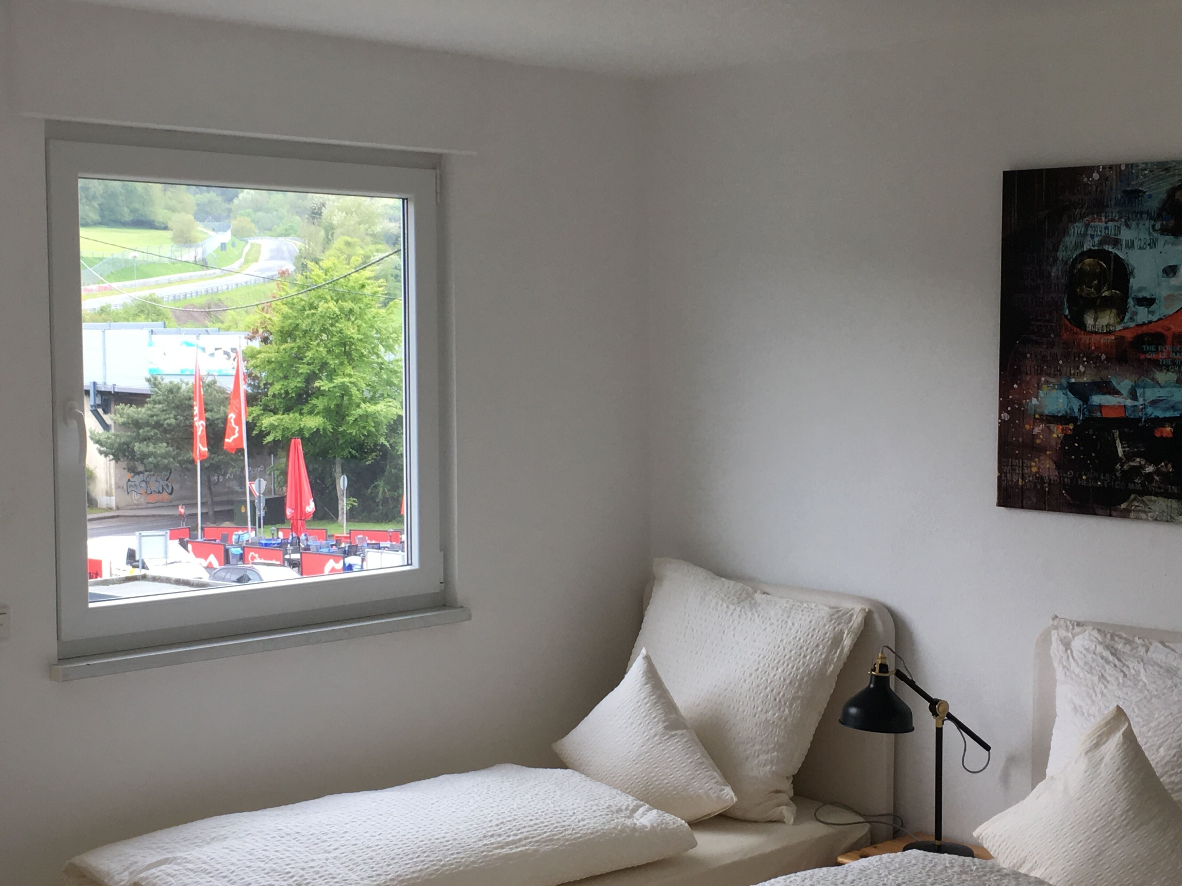 A bedroom with a bed, a bedside lamp, and a window with a view of a street and greenery.