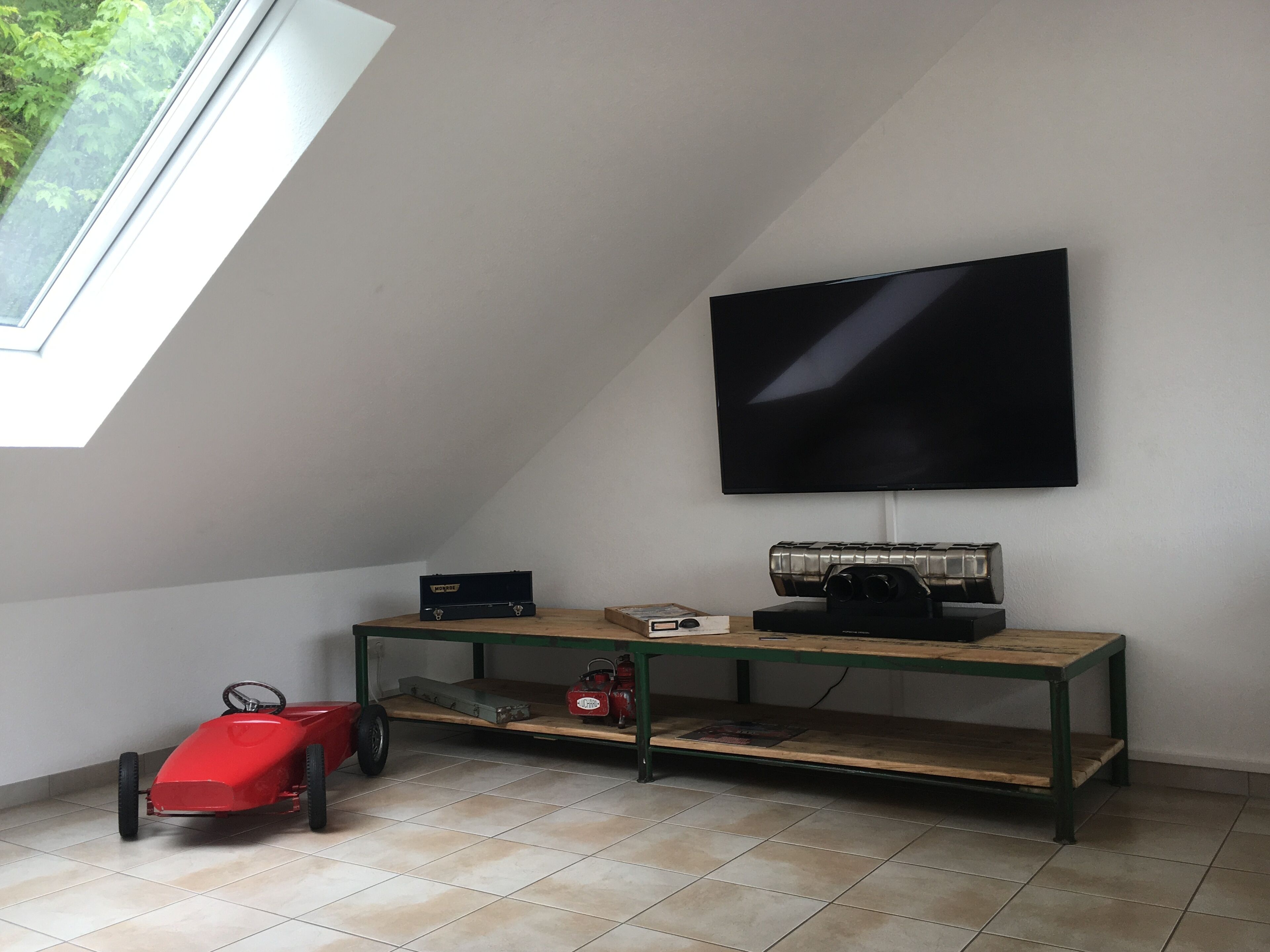 A room with a sloped ceiling, a television mounted on the wall, a wooden table with electronic devices, and a red toy car.