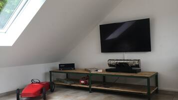 A room with a sloped ceiling, a television mounted on the wall, a wooden table with electronic devices, and a red toy car.