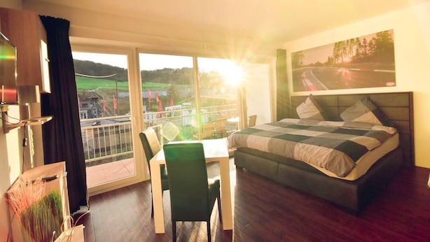 A hotel room with a large bed, a dining table with chairs, a balcony with a view of the countryside, and a painting on the wall. - Nringrooms Exit-Breidscheid (Adenau)