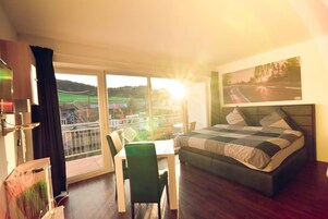 Una habitación de hotel con una cama grande, una mesa de comedor con sillas, un balcón con vistas al campo y un cuadro en la pared.
