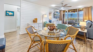 A modern living room with a round glass table, wicker chairs, a grey sofa, and a view of the ocean through large windows. - Amazing Ocean/Beach Front, Pool Side, Ground Floor Remodeled 2 Bdr 2 Bath Condo  (Kihei)