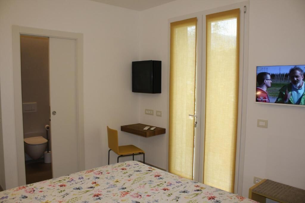 Ein schlichter, kleiner Raum mit einem Bett, einem Sessel, einem Schreibtisch, einem Fernseher und einer Tür zu einem Badezimmer.