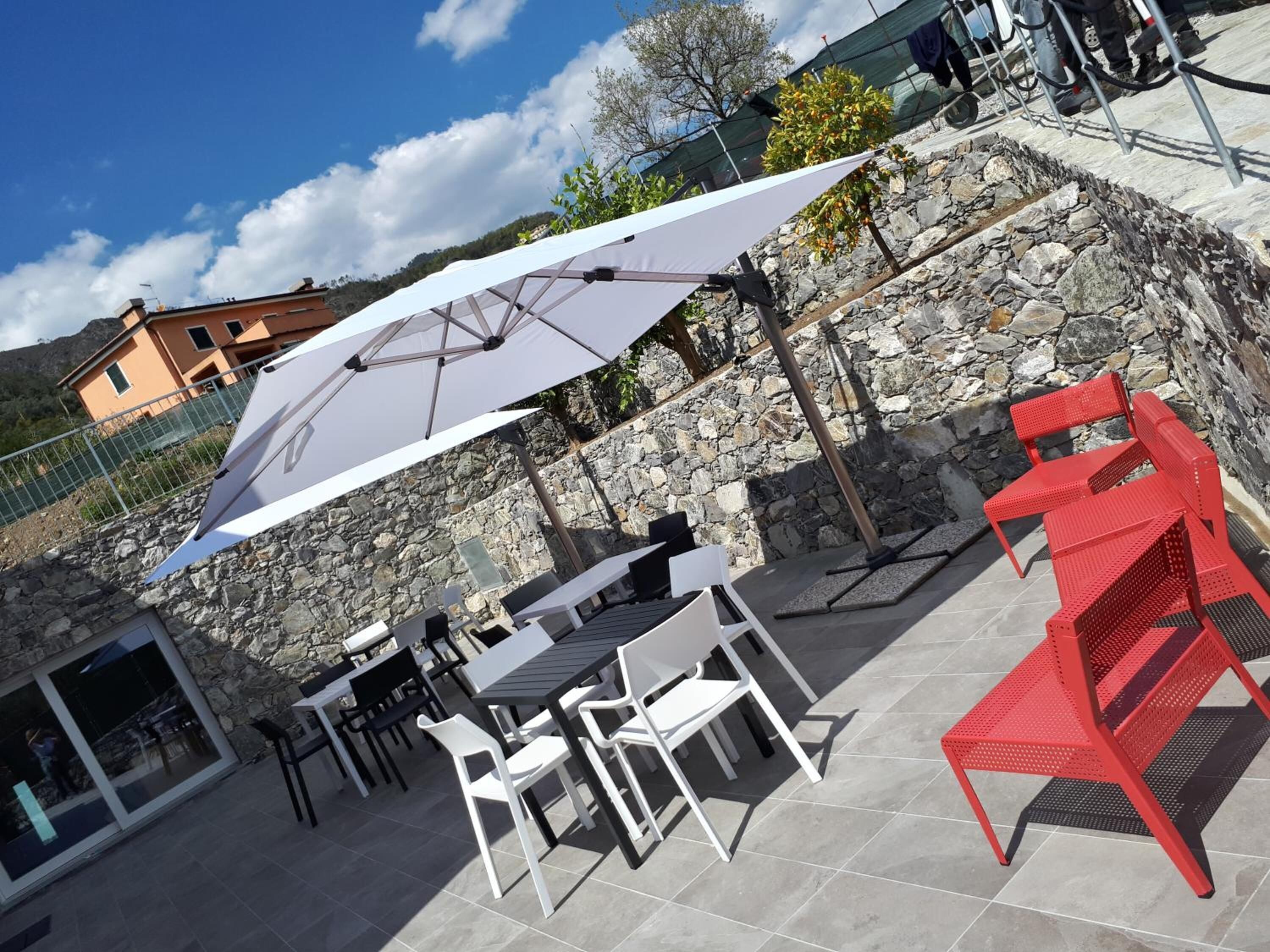 Patio esterno con muri in pietra, un ombrellone bianco e sedie rosse.
