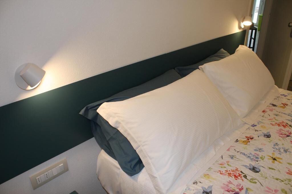 Una cama bien hecha con ropa de cama blanca y gris, un cabecero verde y una lámpara fijada a la pared.