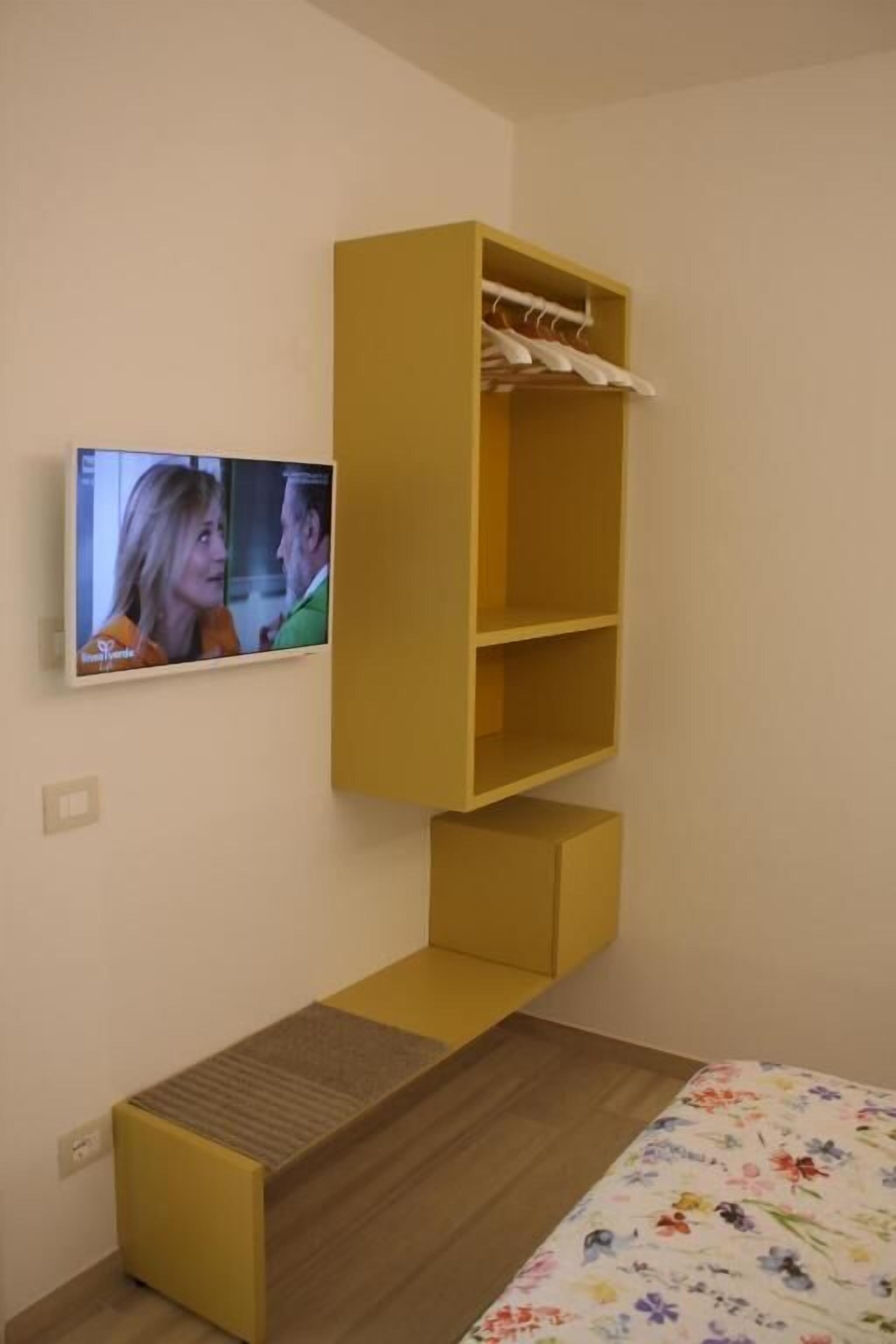 Ein Zimmer mit einem Bett, einem gelben Kleiderschrank, einem an der Wand befestigten Fernseher und einer kleinen Bank.