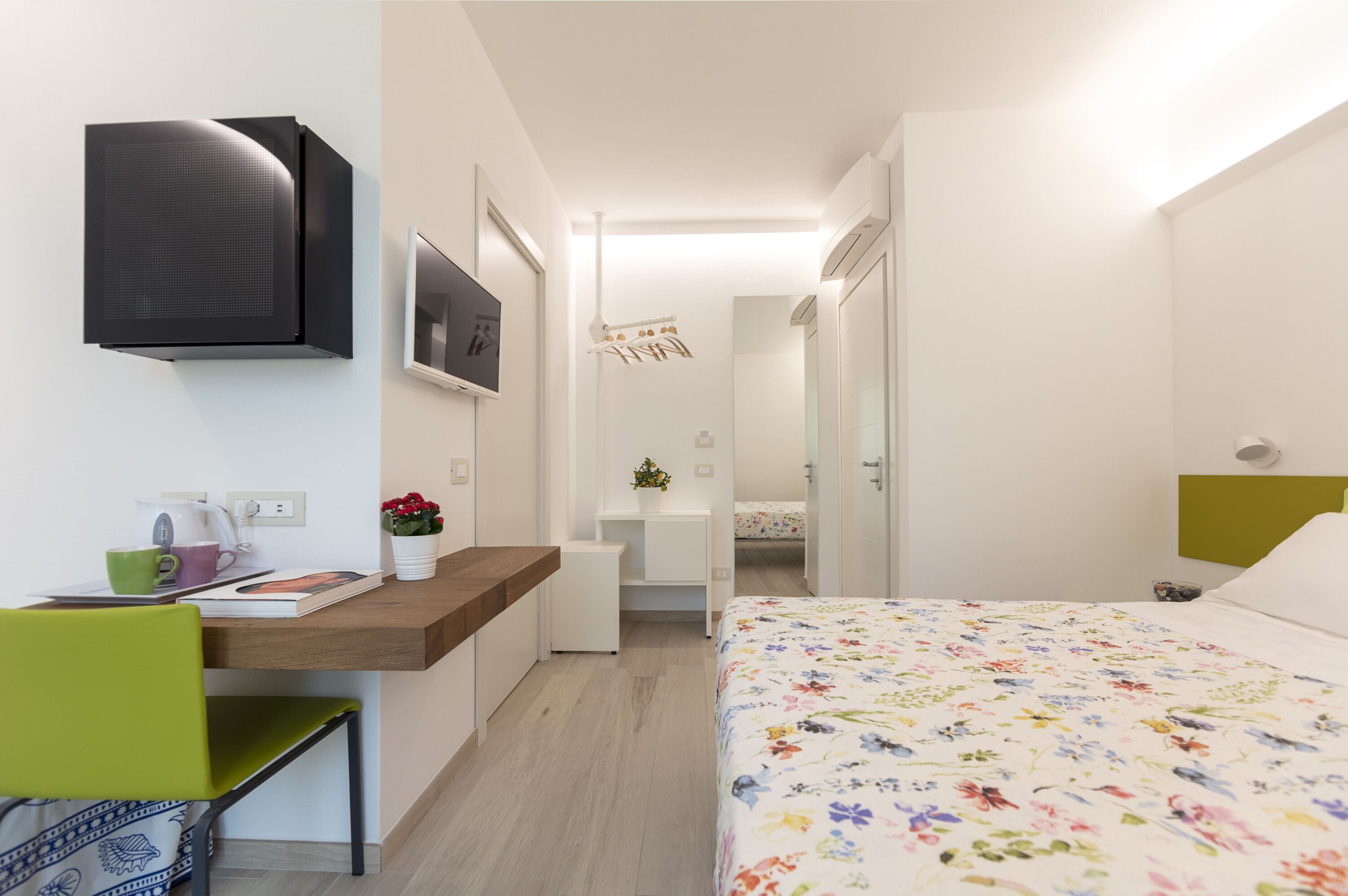 Une chambre d’hôtel moderne avec un bureau en bois, une chaise verte, un lit recouvert d’une courtepointe à motifs floraux, un téléviseur fixé au mur et une petite table sur laquelle est posée une plante.