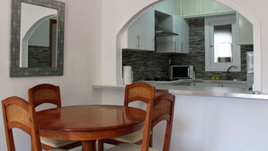 Un comedor con una mesa redonda de madera y cuatro sillas, un espejo grande y una ventana arqueada que da a una cocina moderna con armarios blancos y un salpicadero de piedra.