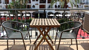Un balcón con mesa de madera y dos sillas de mimbre, con vista a un patio con mobiliario exterior y palmeras.