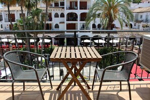 A balcony with a wooden table and two wicker chairs, overlooking a courtyard with outdoor seating and palm trees.