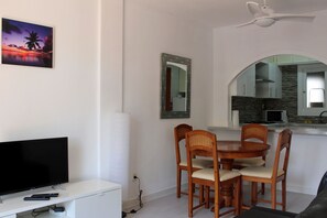 Un coin repas avec une table ronde et quatre chaises, un téléviseur posé sur un meuble blanc, un ventilateur de plafond et une cuisine visible à travers une arche.