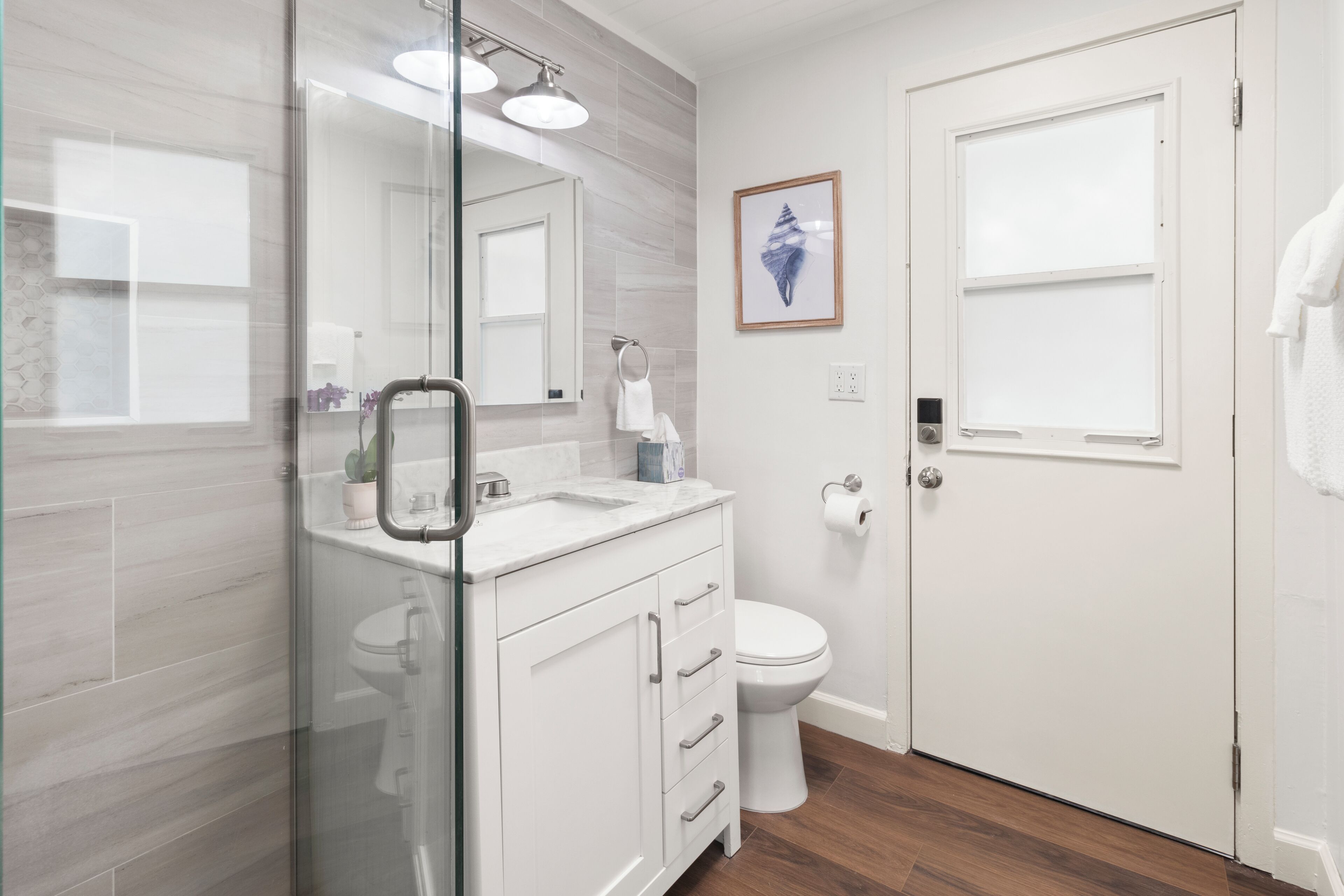 A modern bathroom with a glass shower enclosure, a white sink with a marble countertop, a toilet, and a framed picture on the wall.