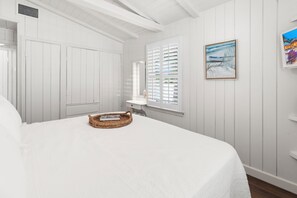 A bedroom with a large bed, white walls, a window with shutters, a painting, and a small bathroom with a sink and mirror. - Cottage Across The Street From #1 Beach (Siesta Key)