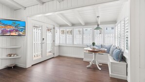 Una habitación con piso de madera, paredes blancas, una mesa redonda con una planta y un televisor a la izquierda.