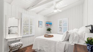 Un dormitorio con cama, tocador con espejo, banca, ventilador de techo y ventana con contraventanas.