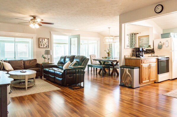 Uma sala de estar espaçosa com sofá de couro, área de jantar com mesa e cadeiras, cozinha com geladeira e cafeteira, e um ventilador de teto.