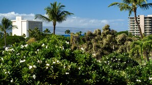 Un exuberante jardín con palmeras y plantas en flor, un edificio moderno con balcones y una vista despejada del océano.