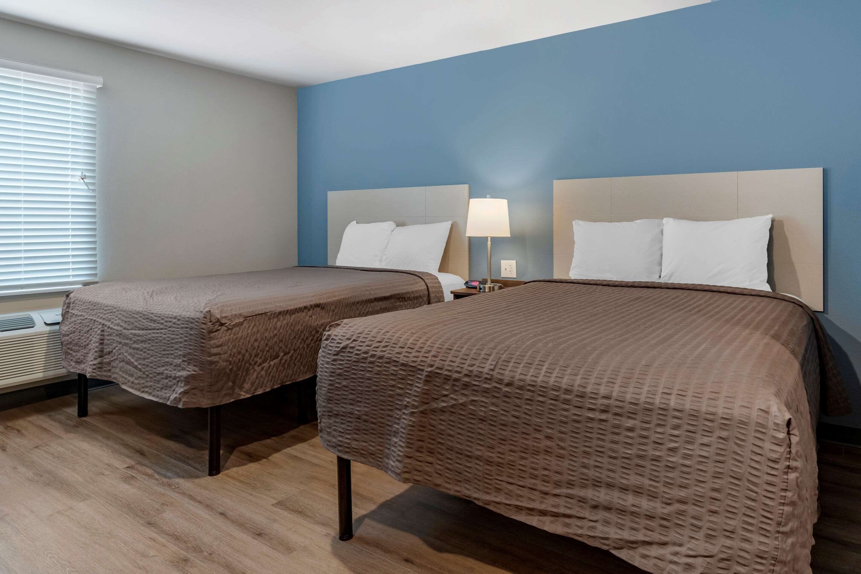 A hotel room with two beds, a window with blinds, a bedside lamp, and a wall heater.
