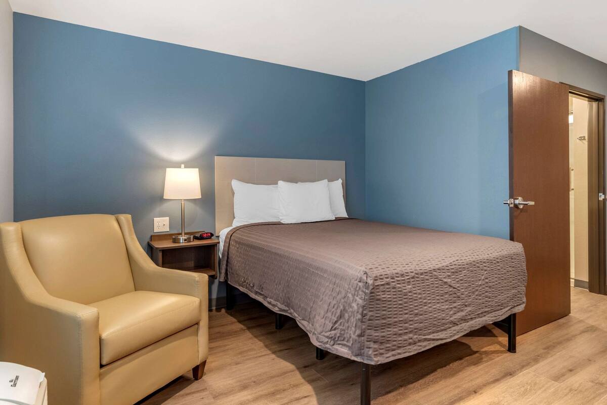 A hotel room with a bed, a chair, a nightstand, and a lamp.