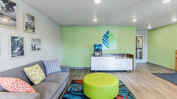 Una sala de estar moderna con un sofá gris, cojines coloridos, un taburete verde y un mueble blanco con una cafetera.
