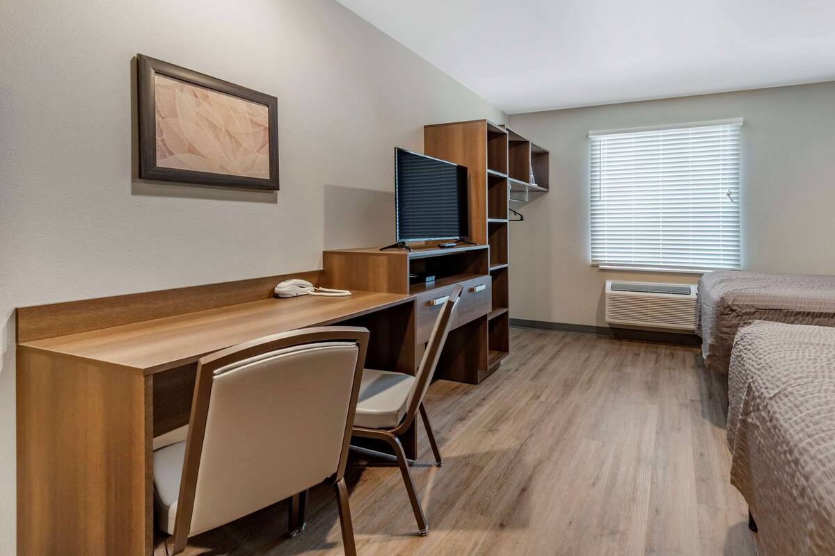 A hotel room with a wooden desk, a chair, a television, a bed, and a window with blinds.