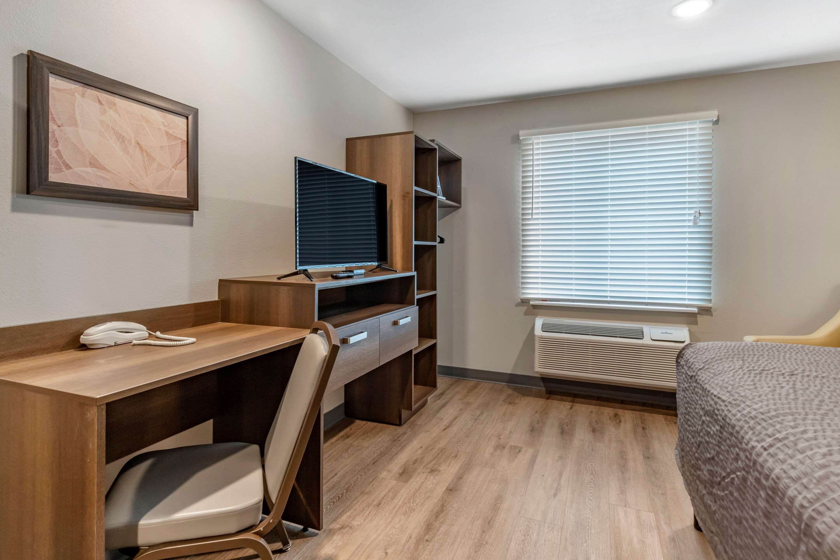 Une chambre d’hôtel avec un bureau en bois, une chaise, une télévision, un lit, une fenêtre avec des stores et un tableau encadré au mur.