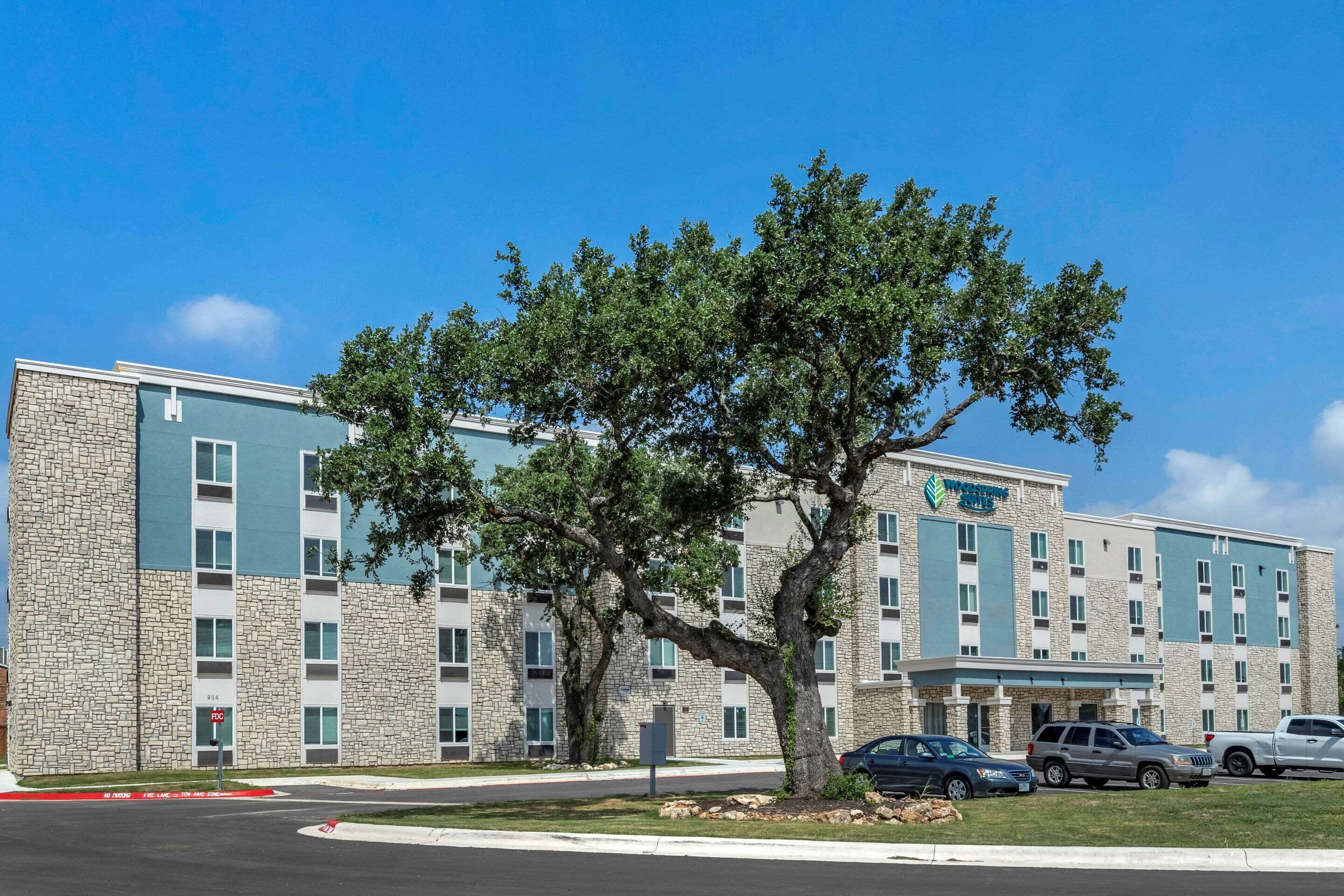 A multi-story hotel with a stone facade, a large tree in front, and several parked cars.