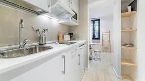 A modern kitchen with white cabinets, stainless steel sink, and a microwave. There is a small dining area with a chair and table. - Apartamentos Madrid Rio (Madrid)