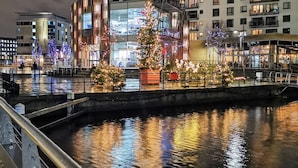 A modern building with a glass facade, a river in front, and Christmas trees with lights. - Modern Leeds City Apartment (Leeds)