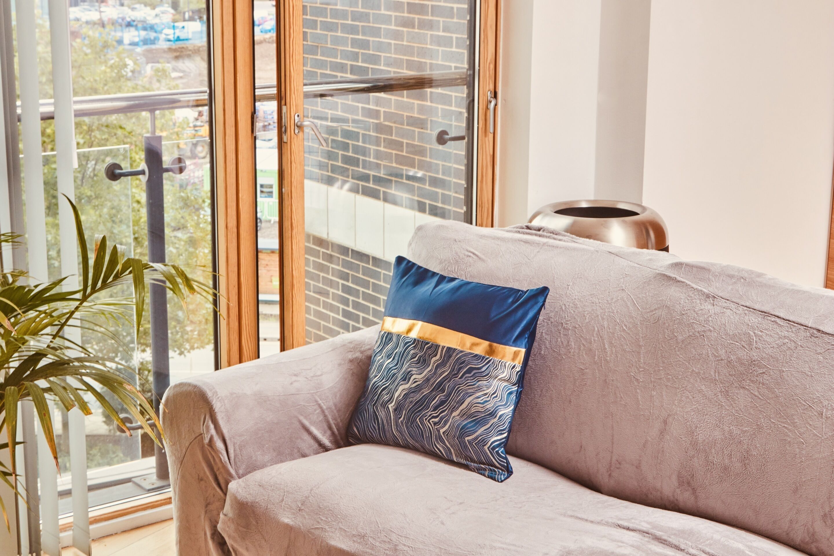 Una habitación con un sofá gris, un cojín estampado, una planta en maceta y vistas a un balcón con barandilla de metal.