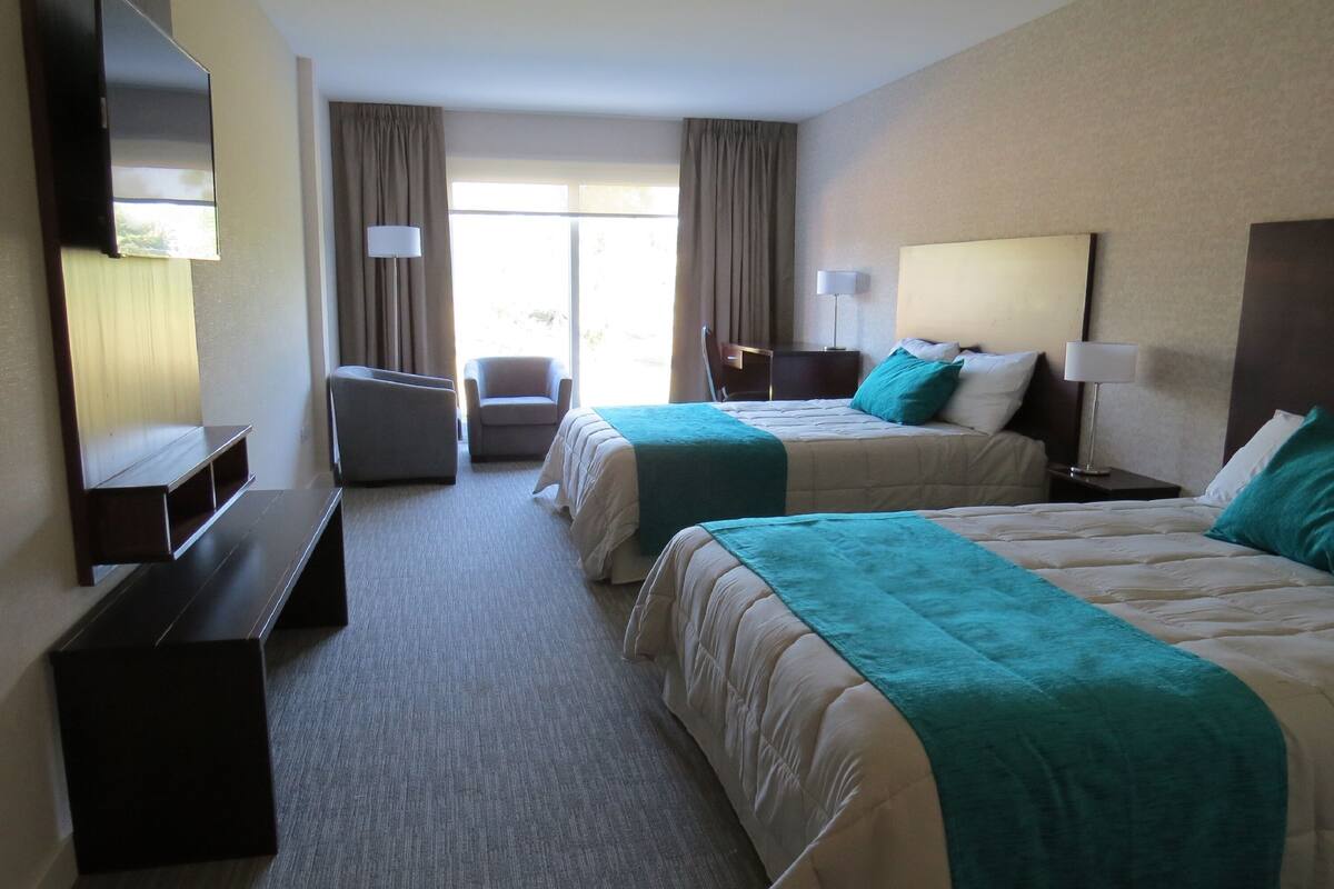 Quarto de hotel com duas camas, uma escrivaninha, uma cadeira, uma televisão e uma janela grande.
