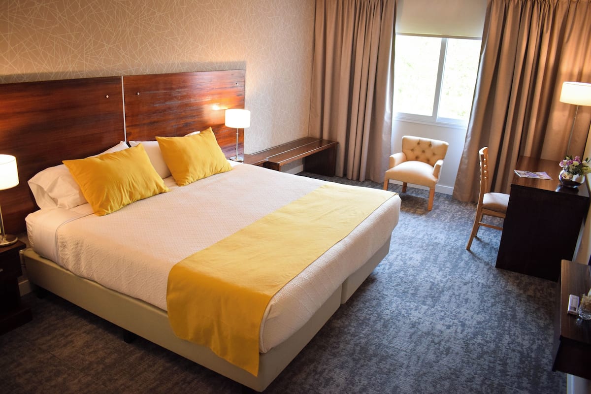 Quarto de hotel com uma cama grande, duas luminárias de cabeceira, uma cadeira, uma escrivaninha e uma janela com cortinas.