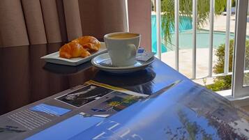 A breakfast setup with croissants and coffee on a table, overlooking a pool and greenery.
