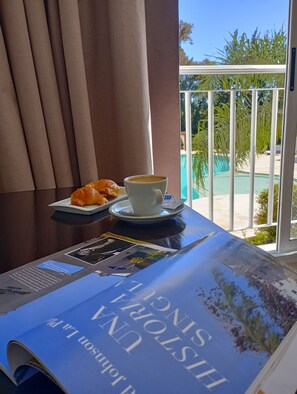 En frukostuppsättning med croissanter och kaffe på ett bord, med utsikt över en pool och grönområden.