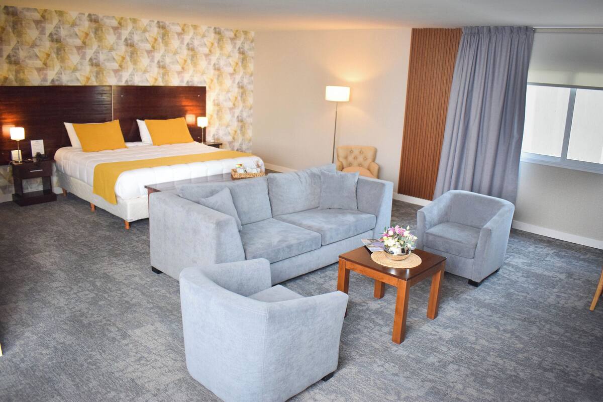 Quarto de hotel com cama, sofá cinza, poltrona e pequena mesa com um vaso de flores.