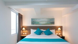 Un lit bien fait, avec une literie blanche et turquoise, deux lampes de chevet et un tableau au-dessus de la tête de lit.