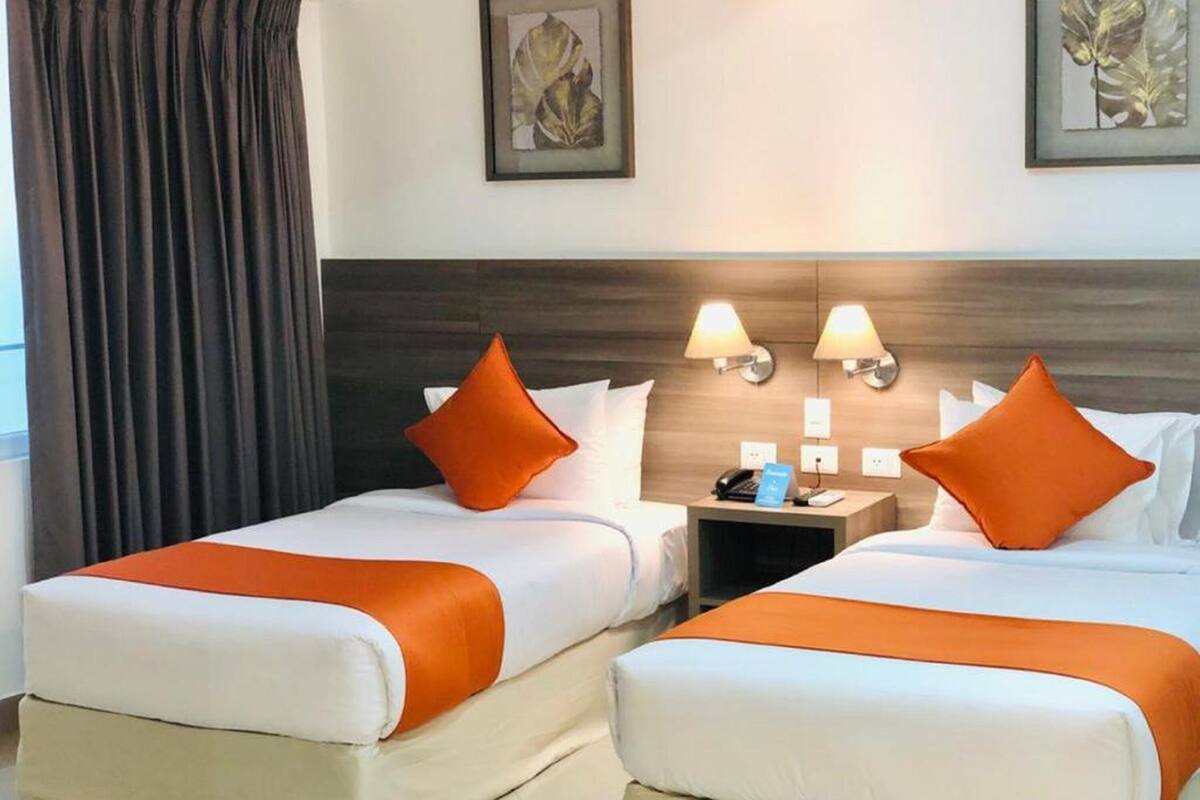 Une chambre d’hôtel avec deux lits, chacun agrémenté d’un coussin orange, dotée de linge de lit blanc et d’une tête de lit en bois. On y trouve deux lampes fixées au mur et une petite table sur laquelle sont posés un téléphone et un panneau.
