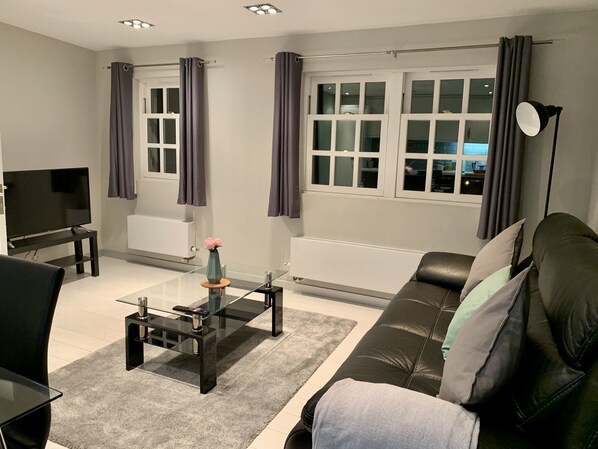 A modern living room with a black leather sofa, a glass coffee table, a TV on a stand, and a window with curtains. - Richmond House by Staytor (Exeter)