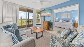 A spacious living room with a sofa, coffee table, and a television. There is a bedroom with a bed and a balcony with curtains. - Remodeled 2BD Condo Across Beach Pool AC - Maui Banyan P301 (Kihei)