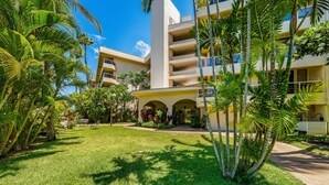 A hotel with a central courtyard, palm trees, and a building with multiple balconies. - Remodeled 2BD Condo Across Beach Pool AC - Maui Banyan P301 (Kihei)