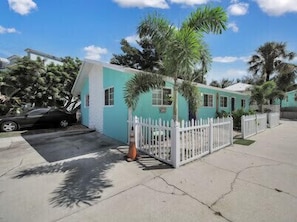 Une maison turquoise avec une clôture blanche en bois, une voiture garée et des palmiers.