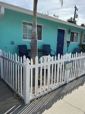 Un bâtiment turquoise avec une clôture blanche en bois, une porte bleue et deux chaises à l’extérieur.