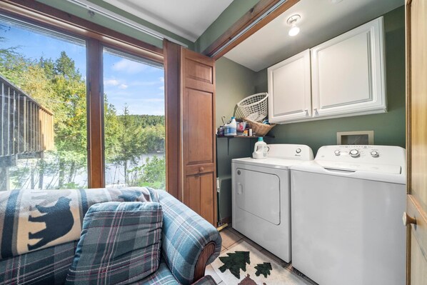 A laundry room with a washing machine and dryer, a wooden door, and a view of trees through a window.
