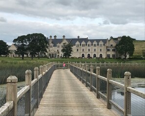 A wooden bridge leading to a large, historic building with multiple chimneys and arched windows. - The Gate Lodge (Ballyclare)